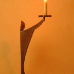「月のあかり」という鉄の燭台。月の形をした頭部を持つ人のカタチに模したロウソク立てで、灯りを差し出すような佇まいをしています。カタチにより、佑・炬・瑛の３シリーズになっており、祐（ゆう）は、両手を差し伸べて救う、炬（きょ）は、松明のように掲げる火、瑛（えい）は、光り輝く玉という意味があります。暗い中で小さな和蝋燭を灯した状態での写真です。頭部の影が月のカタチに揺れて、個体によってはスリットから漏れる光が日輪となったり、または三日月となったりします。