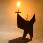 「月のあかり」という鉄の燭台。月の形をした頭部を持つ人のカタチに模したロウソク立てで、灯りを差し出すような佇まいをしています。カタチにより、佑・炬・瑛の３シリーズになっており、祐（ゆう）は、両手を差し伸べて救う、炬（きょ）は、松明のように掲げる火、瑛（えい）は、光り輝く玉という意味があります。暗い中で小さな和蝋燭を灯した状態での写真です。頭部の影が月のカタチに揺れて、個体によってはスリットから漏れる光が日輪となったり、または三日月となったりします。