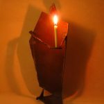 「月のあかり」という鉄の燭台。月の形をした頭部を持つ人のカタチに模したロウソク立てで、灯りを差し出すような佇まいをしています。カタチにより、佑・炬・瑛の３シリーズになっており、祐（ゆう）は、両手を差し伸べて救う、炬（きょ）は、松明のように掲げる火、瑛（えい）は、光り輝く玉という意味があります。暗い中で小さな和蝋燭を灯した状態での写真です。頭部の影が月のカタチに揺れて、個体によってはスリットから漏れる光が日輪となったり、または三日月となったりします。