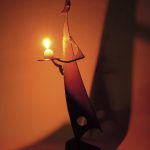 「月のあかり」という鉄の燭台。月の形をした頭部を持つ人のカタチに模したロウソク立てで、灯りを差し出すような佇まいをしています。カタチにより、佑・炬・瑛の３シリーズになっており、祐（ゆう）は、両手を差し伸べて救う、炬（きょ）は、松明のように掲げる火、瑛（えい）は、光り輝く玉という意味があります。暗い中で小さな和蝋燭を灯した状態での写真です。頭部の影が月のカタチに揺れて、個体によってはスリットから漏れる光が日輪となったり、または三日月となったりします。
