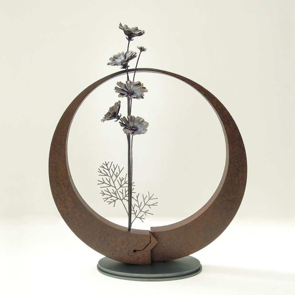 円の鉄プレートに鍛鉄のコスモスの花が添えられたアート作品です。 中心をずらした円でくり抜かれた円形プレートは重厚な錆仕上げ。タイトに纏めたコスモスの花束は上品な半つやで鉄地金の色味を生かした表現です。鍛造、溶接で変化した鉄の色味が見えています。 花は蕾から咲き誇る状態まで変化のある表現で、デザイン化された葉は平面的にアレンジし足元を装飾しています。