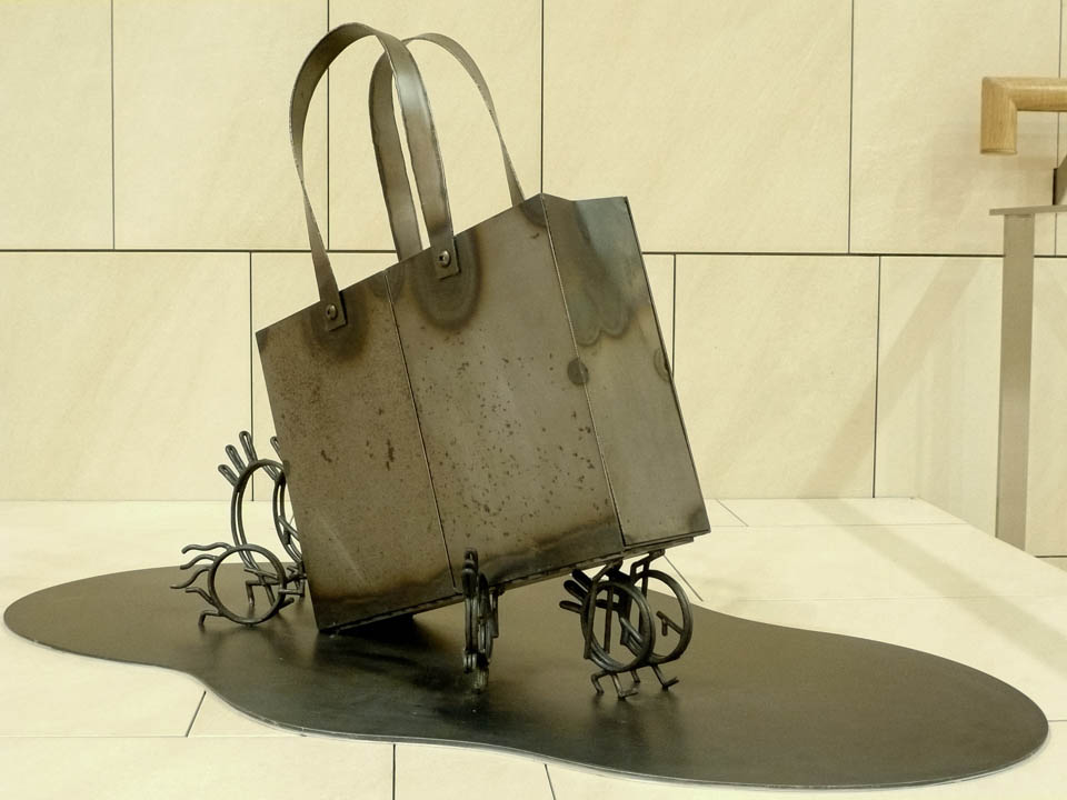 横浜ノースポートモールにオープン時設置されていたアートオブジェ「急ぐヤツら」。忘れ物のバッグや鞄を届けるために駅へと急いでいる彫刻作品です。全て鉄で制作されました。トートバッグを運ぶ作品。