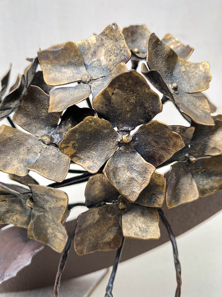 紫陽花のこんもりした花が円の中に添えられているアート作品、鉄の彫刻です。柔らかい花びらを模した鉄のアジサイの花に、面と線で表現した葉が寄り添っています。花のアップ写真。