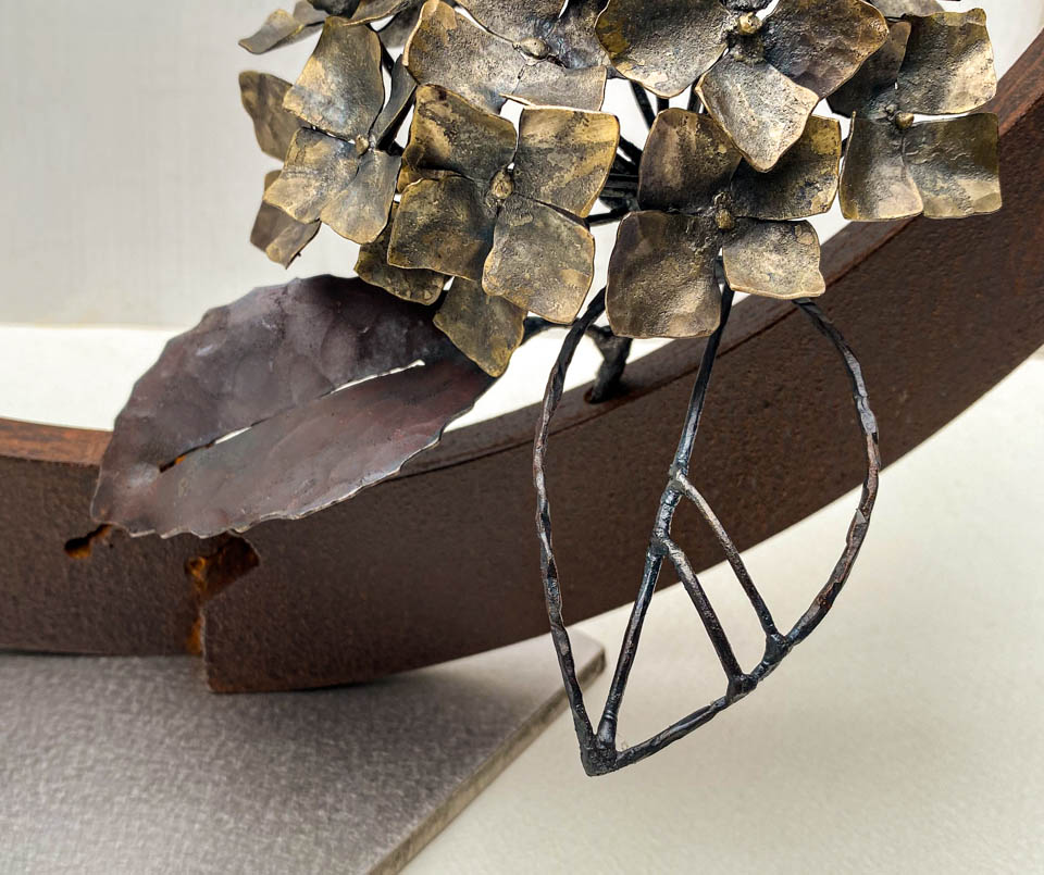 紫陽花のこんもりした花が円の中に添えられているアート作品、鉄の彫刻です。柔らかい花びらを模した鉄のアジサイの花に、面と線で表現した葉が寄り添っています。葉のアップ写真。