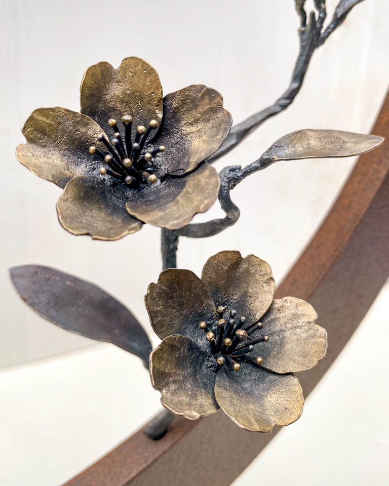 箱根に咲くマメザクラの小さな枝が、丸いベースに添えられた鉄の彫刻です。かわいい花が鉄で繊細に表現され、新芽が芽吹いています。花の表現。