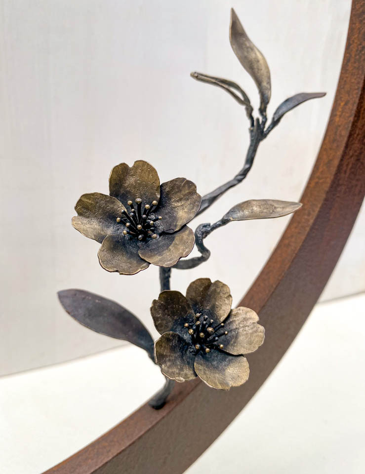箱根に咲くマメザクラの小さな枝が、丸いベースに添えられた鉄の彫刻です。かわいい花が鉄で繊細に表現され、新芽が芽吹いています。花の表現。
