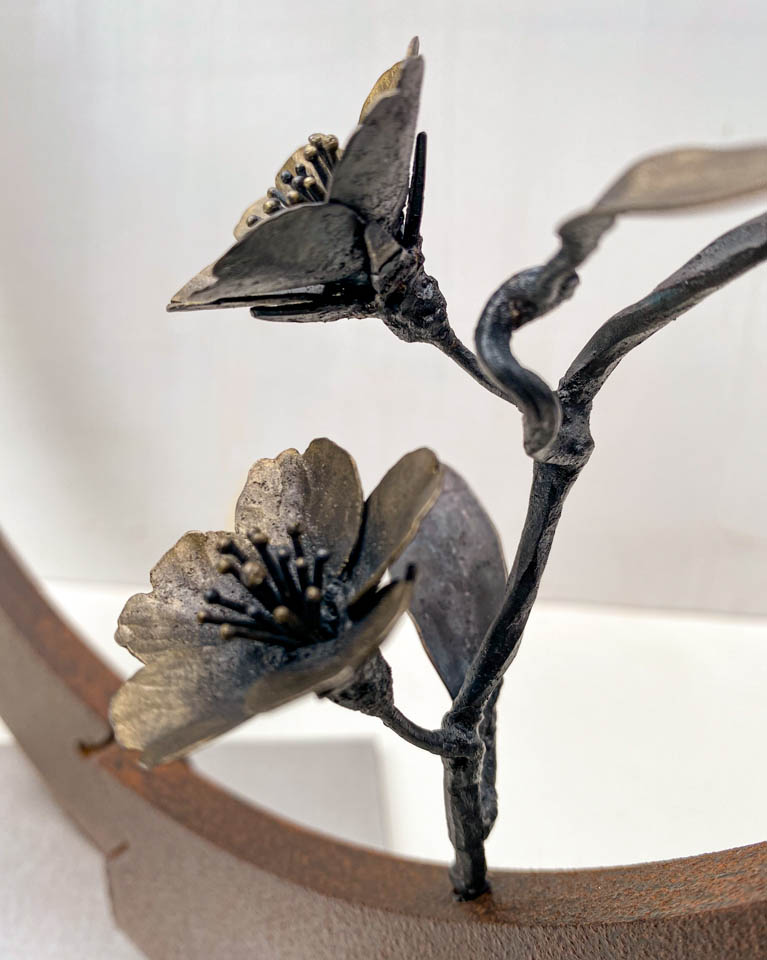 箱根に咲くマメザクラの小さな枝が、丸いベースに添えられた鉄の彫刻です。かわいい花が鉄で繊細に表現され、新芽が芽吹いています。花の表現。