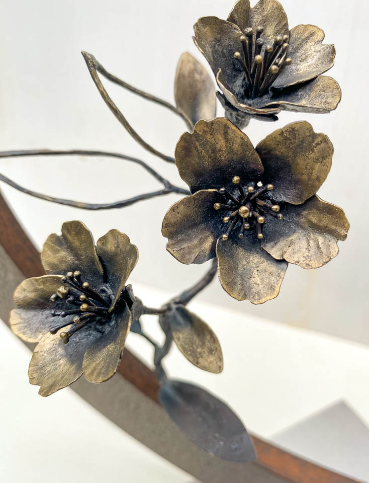 箱根に咲くマメザクラの小さな枝が、丸いベースに添えられた鉄の彫刻です。かわいい花が鉄で繊細に表現され、新芽が芽吹いています。花の表現。