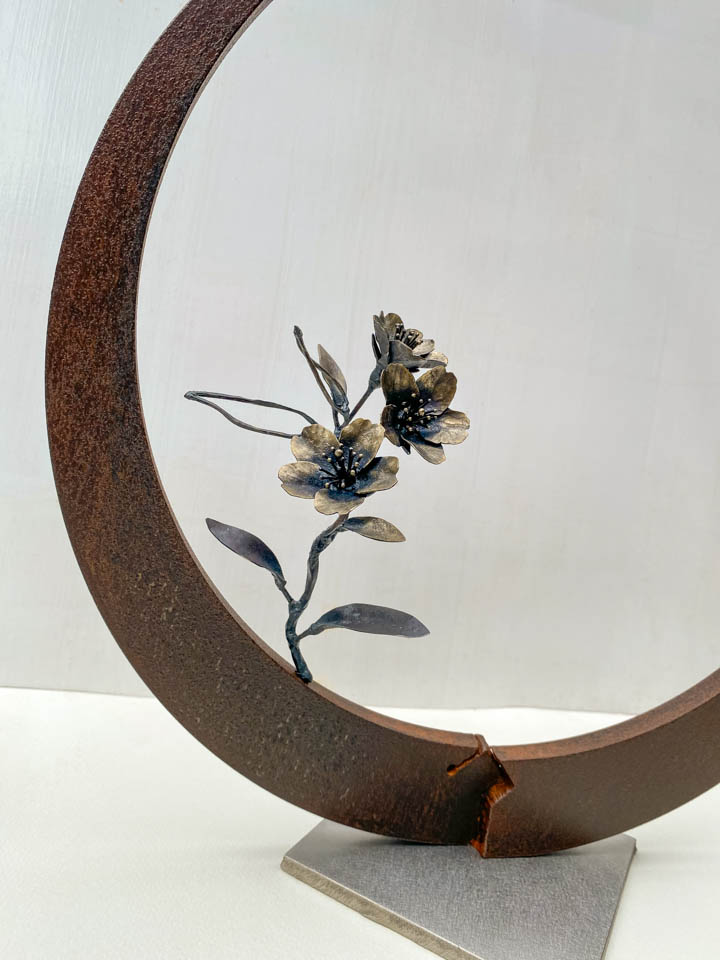 箱根に咲くマメザクラの小さな枝が、丸いベースに添えられた鉄の彫刻です。かわいい花が鉄で繊細に表現され、新芽が芽吹いています。花の表現。