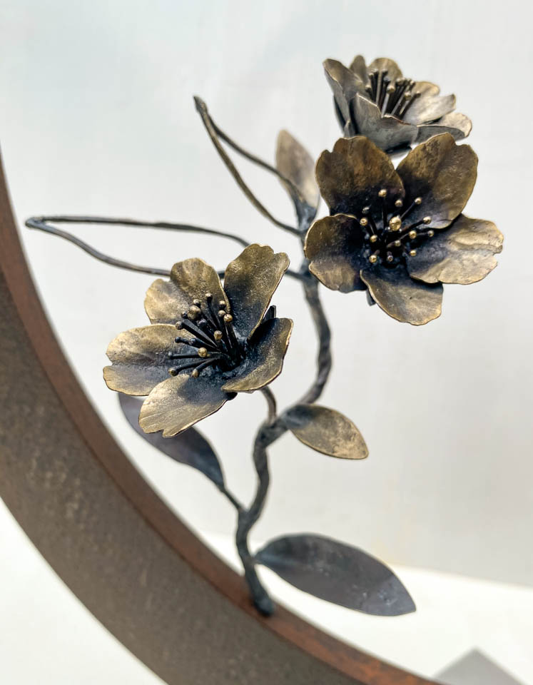 箱根に咲くマメザクラの小さな枝が、丸いベースに添えられた鉄の彫刻です。かわいい花が鉄で繊細に表現され、新芽が芽吹いています。花の表現。