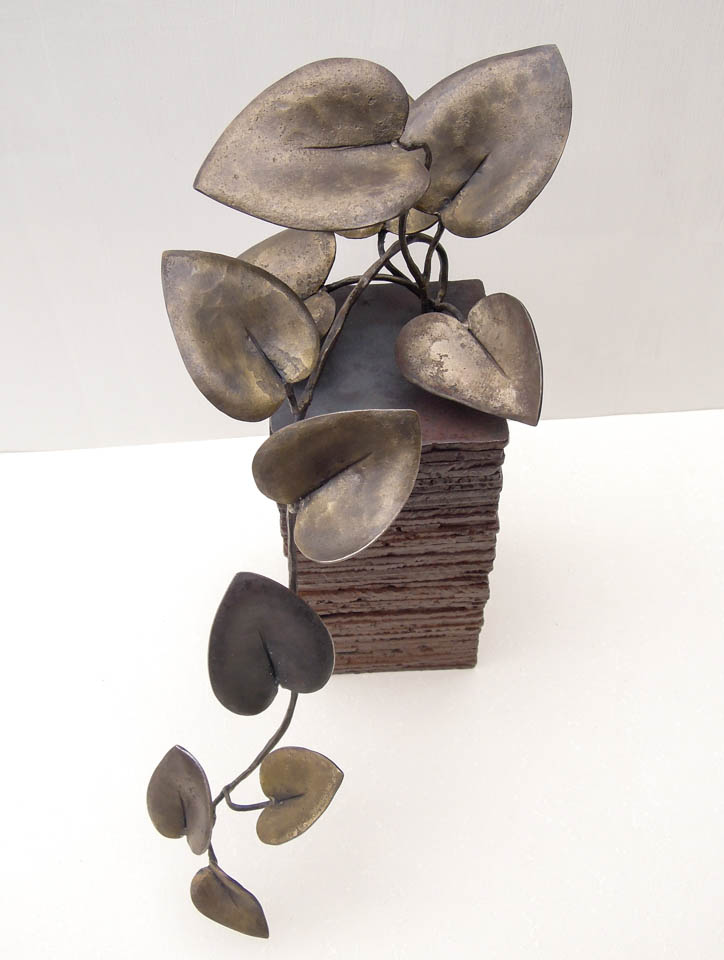 蔓(カズラ)をモチーフにした鉄の彫刻、アートオブジェです。 ツルが上から下へ垂れながら葉を光に向かって持ち上げる様子を、躍動感ある姿として表現しました。 一枚はアクセントとして黒みのある仕上がりとし、他は真鍮ブラシで磨き込み黄色みを帯びた控えめが光沢があります。 厚みのある鉄板を重ねたベースからツルがこぼれ出ている姿です。 葉の表情。