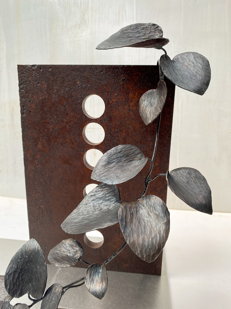 丸い開口が連なる表情豊かな鉄の厚板に、カンガルーアイビーのツルが肩から下がるよう配置した鉄のアート作品です。華やかな大小の葉がリズミカルに連なり、軽やかな印象の彫刻作品になっています。部分写真。